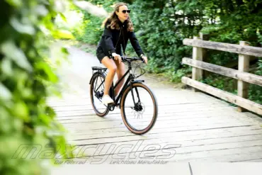 Eine Frau mit Sonnenbrille und schwarzer Jacke fährt auf einem schwarzen Steel Lite 2 Trapez Fahrrad auf einer Holzbrücke, umgeben von Bäumen.