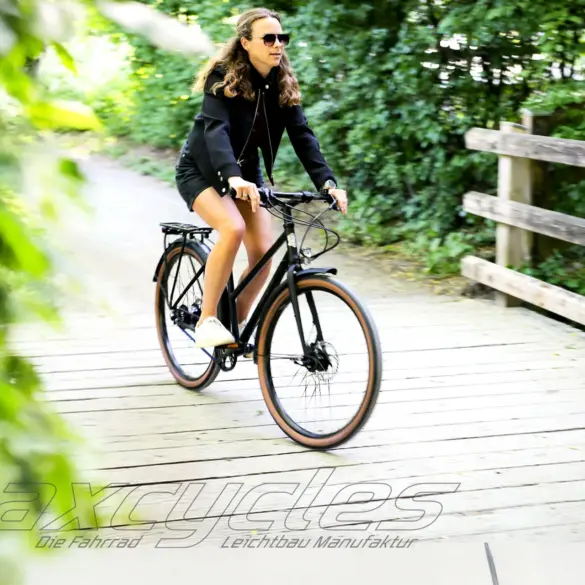 Eine Frau mit Sonnenbrille und schwarzer Jacke fährt auf einem schwarzen Steel Lite 2 Trapez Fahrrad auf einer Holzbrücke, umgeben von Bäumen.