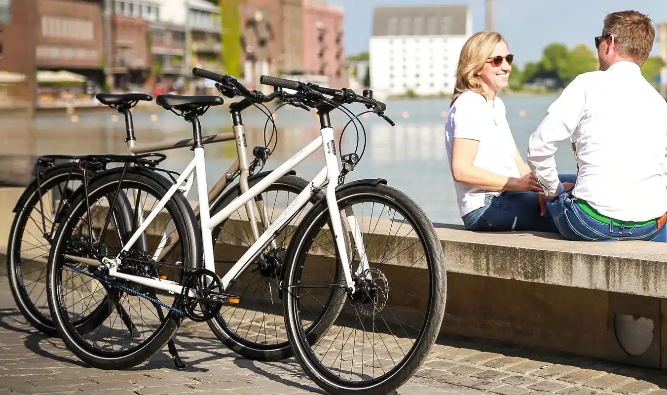 Zwei Fahrräder, ein weißes und ein hellgraues, des Modells Steel Lite 2 Trapez von Maxcycles stehen nebeneinander im Freien. Im Hintergrund sitzen ein Mann und eine Frau auf einer Mauer am Wasser.