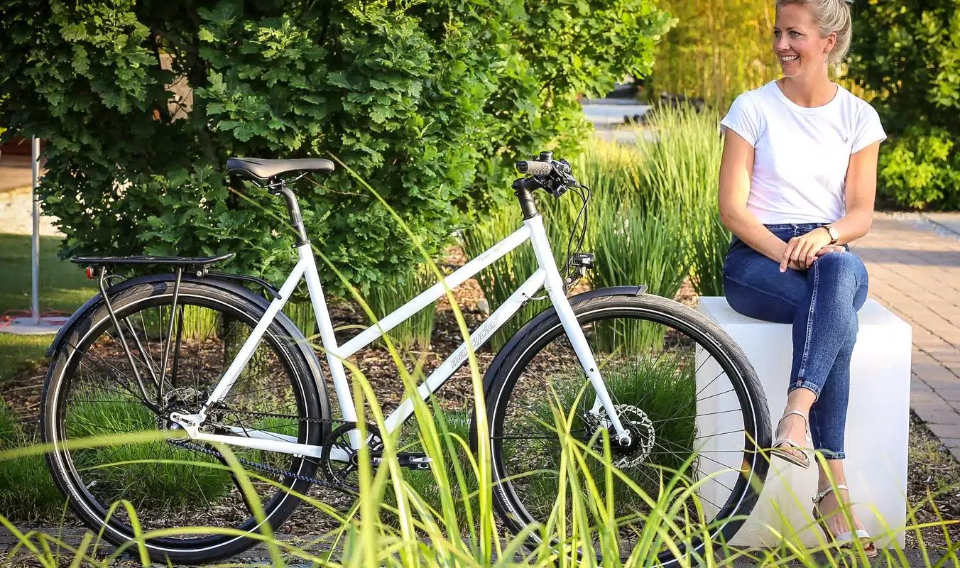 Eine Frau sitzt lächelnd neben einem weißen Steel Lite 2 Trapez Fahrrad, das mit einem Gepäckträger und Schutzblechen ausgestattet ist, im Freien.
