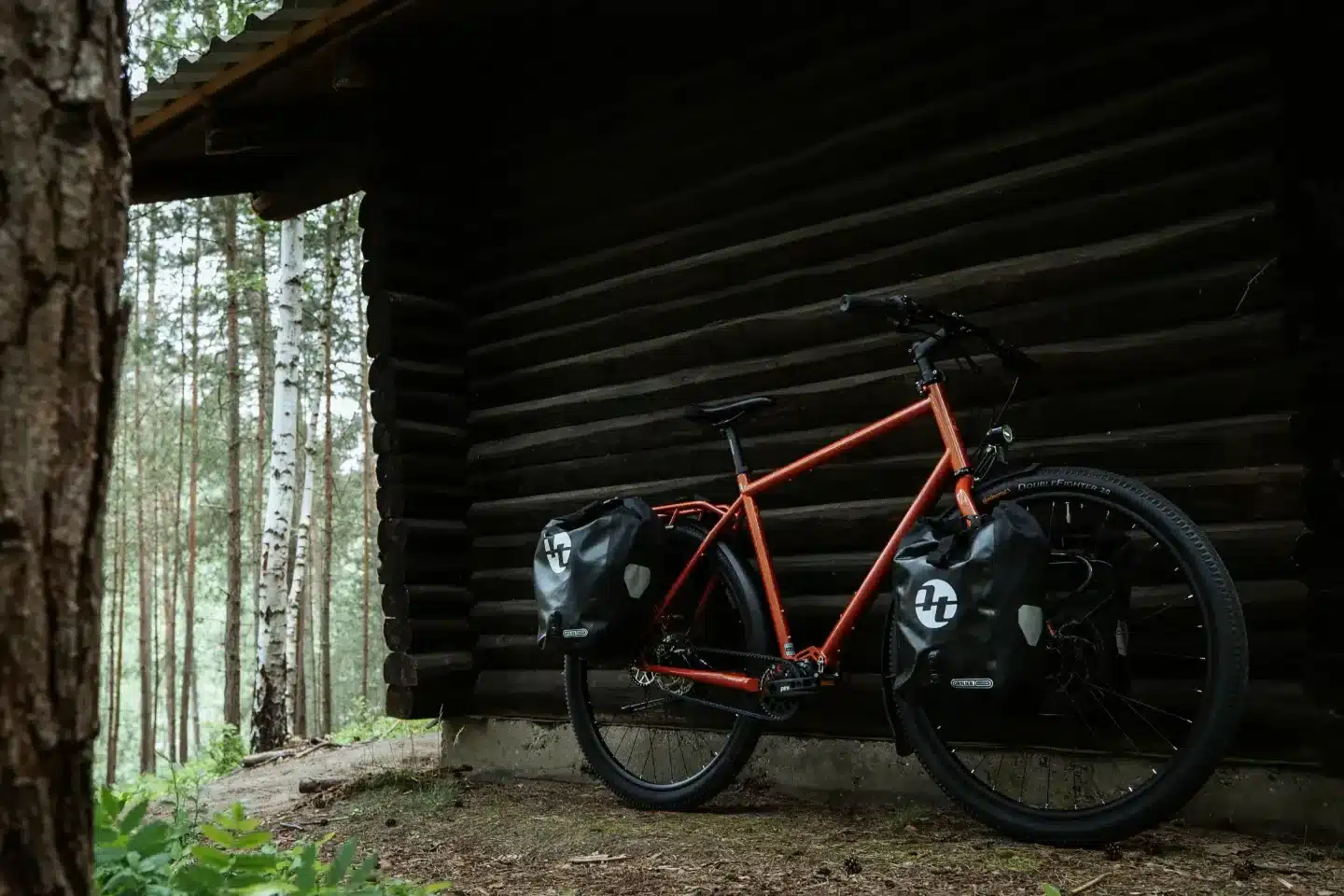 Ein oranges Tout Terrain Tanami Fahrrad mit Packtaschen, abgestellt vor einer Holzhütte im Wald.