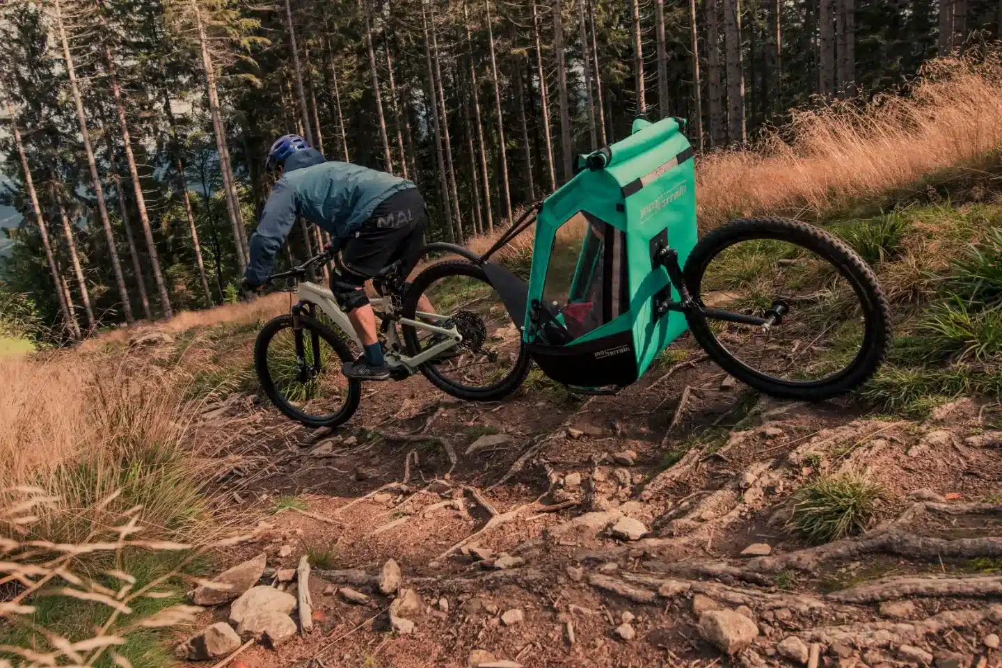 Mountainbiker fährt mit einem grünen Tout Terrain Singletrailer II Sport Kinderanhänger auf einem schmalen, wurzeligen Waldpfad bergab.