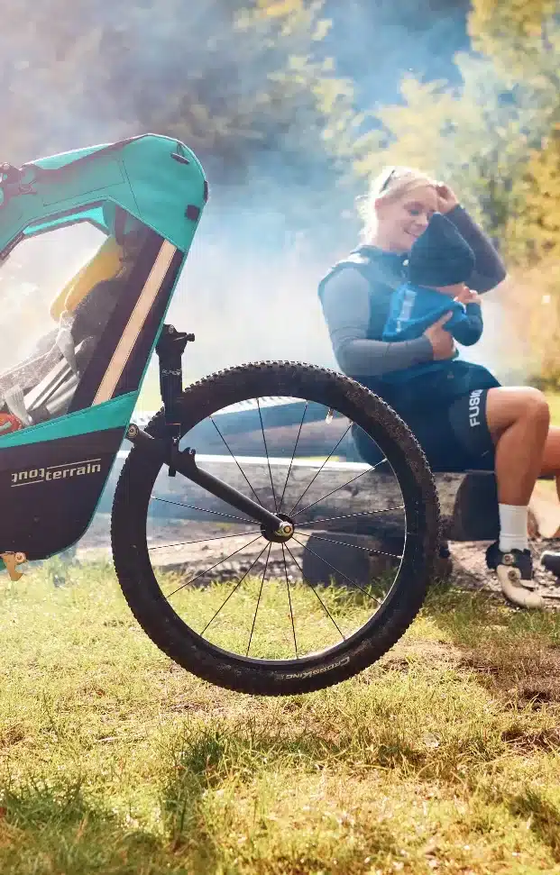 Ein Tout Terrain Singletrailer II Sport Kinderanhänger mit 24-Zoll-Rad steht im Gras, im Hintergrund eine Frau mit Kind auf einer Holzbank.
