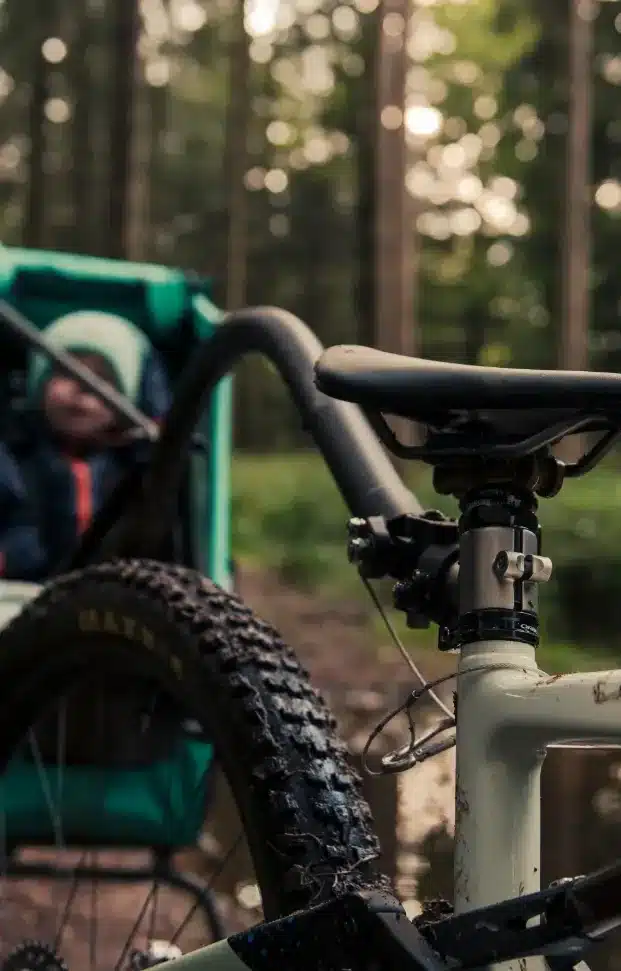 Detailaufnahme eines Tout Terrain Singletrailer II Sport Kinderanhängers, befestigt an einem Mountainbike, mit einem Kind im Hintergrund und Waldbäumen.
