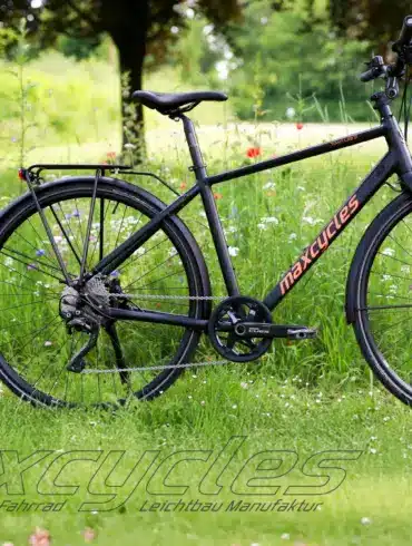 Schwarzes Maxcycles Town Lite 2 Cross Fahrrad steht auf einer grünen Wiese mit Blumen.