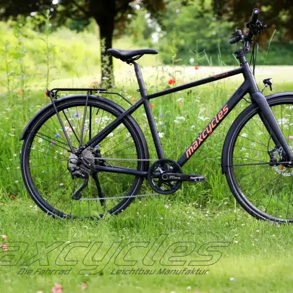 Schwarzes Maxcycles Town Lite 2 Cross Fahrrad steht auf einer grünen Wiese mit Blumen.