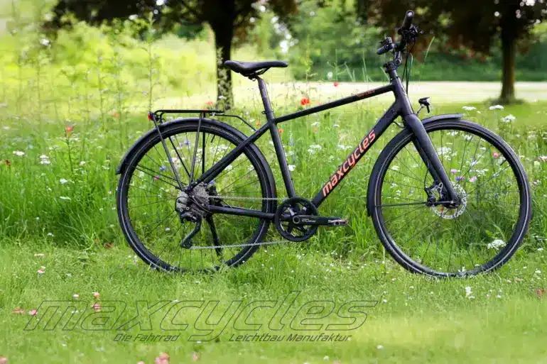 Schwarzes Maxcycles Town Lite 2 Cross Fahrrad steht auf einer grünen Wiese mit Blumen.