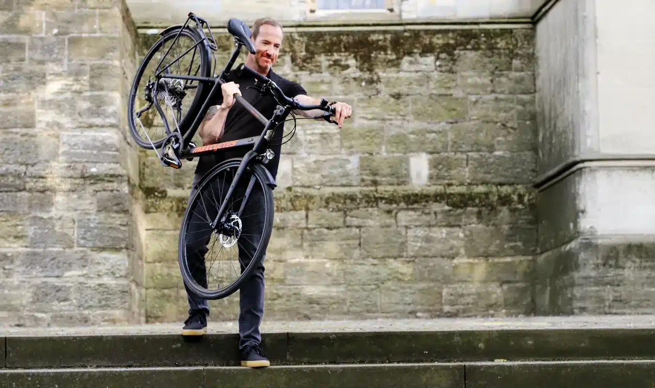 Ein Mann mit Bart trägt ein schwarzes Town Lite 2 Cross Fahrrad, das mit einem Gepäckträger ausgestattet ist.