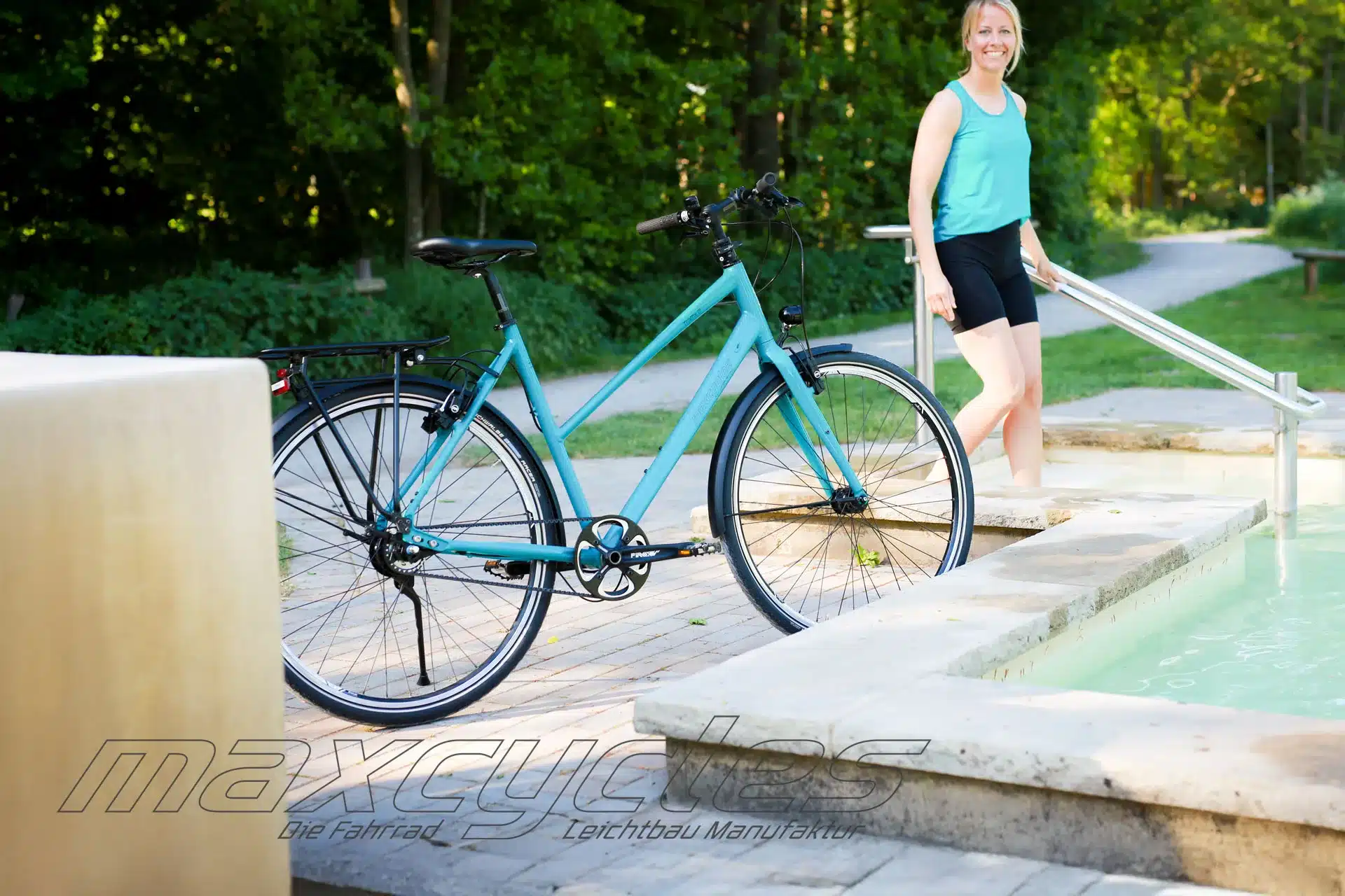Türkises Stadtrad der Marke Maxcycles mit Gepäckträger und Schutzblechen steht neben einer Frau, die in ein Wasserbecken steigt.