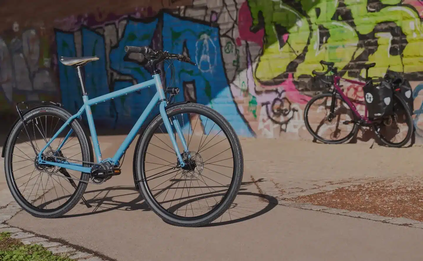 Hellblaues Tout Terrain Tribeca Fahrrad mit flachem Lenker, das auf einem Weg vor einer bunten Graffiti-Wand steht. Ein weiteres Fahrrad ist im Hintergrund zu sehen.