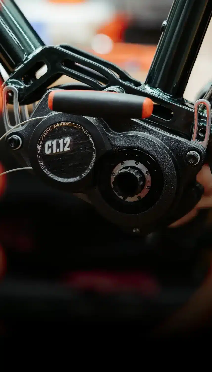 Detailansicht des schwarzen Pinion C1.12 Schaltgetriebes mit orangefarbenen Akzenten, montiert an einem dunkelgrünen Fahrradrahmen, mit Fokus auf die Technologie für wartungsarmen Antrieb.