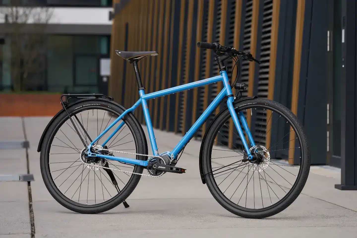 Seitenansicht eines blauen Tout Terrain Tribeca Fahrrads mit Flachlenker, Nabenschaltung und Gepäckträgern, abgestellt auf einem Gehweg vor einer modernen Holzfassade.