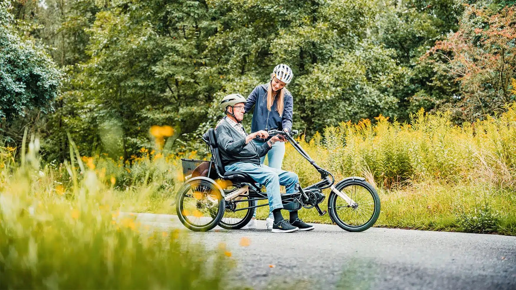 Ein älterer Herr mit Helm sitzt auf einem Trigo Dreirad im Freien, eine junge Frau mit Helm steht neben ihm und lächelt.