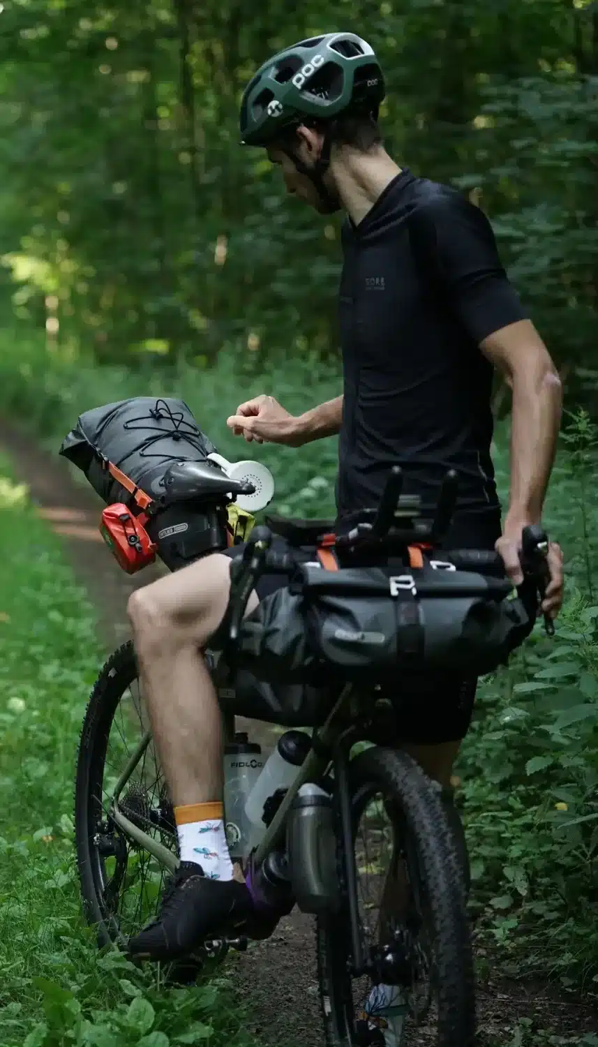 Ein Radfahrer in voller Montur steht mit seinem mit Gepäcktaschen beladenen Bikepacking-Fahrrad auf einem Waldweg.