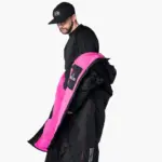 Mann mit schwarzer Kappe und schwarzem Oberteil, der eine schwarze, übergroße Robe mit leuchtend pinkem Teddy-Fleece-Futter präsentiert. Die Robe wird seitlich geöffnet gezeigt, um das Innenfutter hervorzuheben.