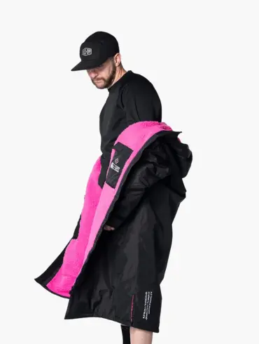 Mann mit schwarzer Kappe und schwarzem Oberteil, der eine schwarze, übergroße Robe mit leuchtend pinkem Teddy-Fleece-Futter präsentiert. Die Robe wird seitlich geöffnet gezeigt, um das Innenfutter hervorzuheben.