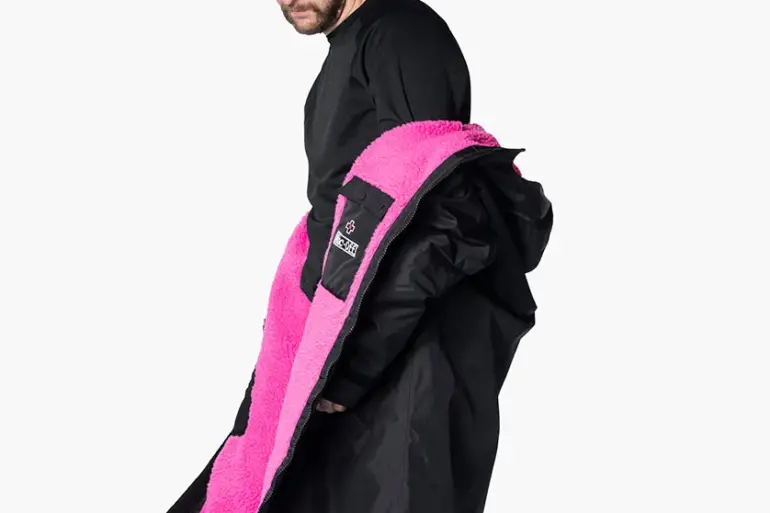 Mann mit schwarzer Kappe und schwarzem Oberteil, der eine schwarze, übergroße Robe mit leuchtend pinkem Teddy-Fleece-Futter präsentiert. Die Robe wird seitlich geöffnet gezeigt, um das Innenfutter hervorzuheben.