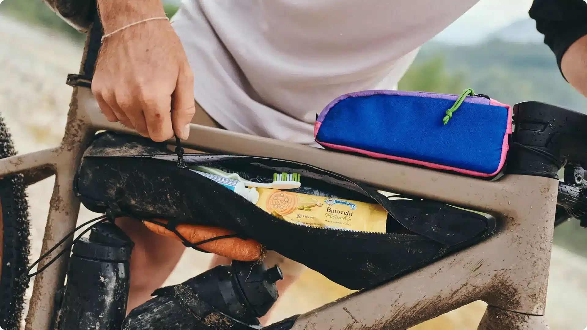 Ein Mann hält eine schwarze Rahmentasche an einem Gravelbike offen, die Zahnbürste, Zahnpasta und Kekse enthält. Daneben liegt eine bunte Kulturtasche.