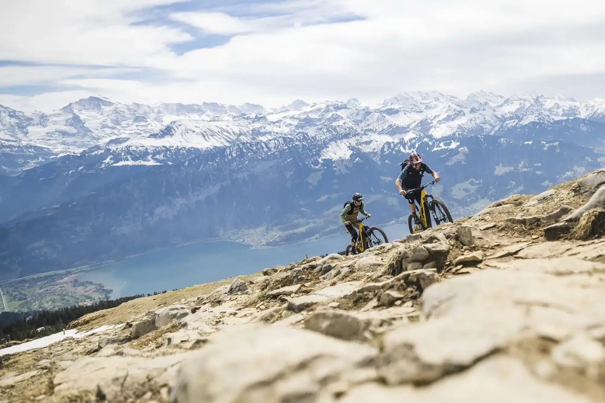 Zwei Mountainbiker, die auf ihren E-Bikes einen felsigen Bergpfad hinunterfahren, mit einer atemberaubenden Berglandschaft und einem See im Tal.