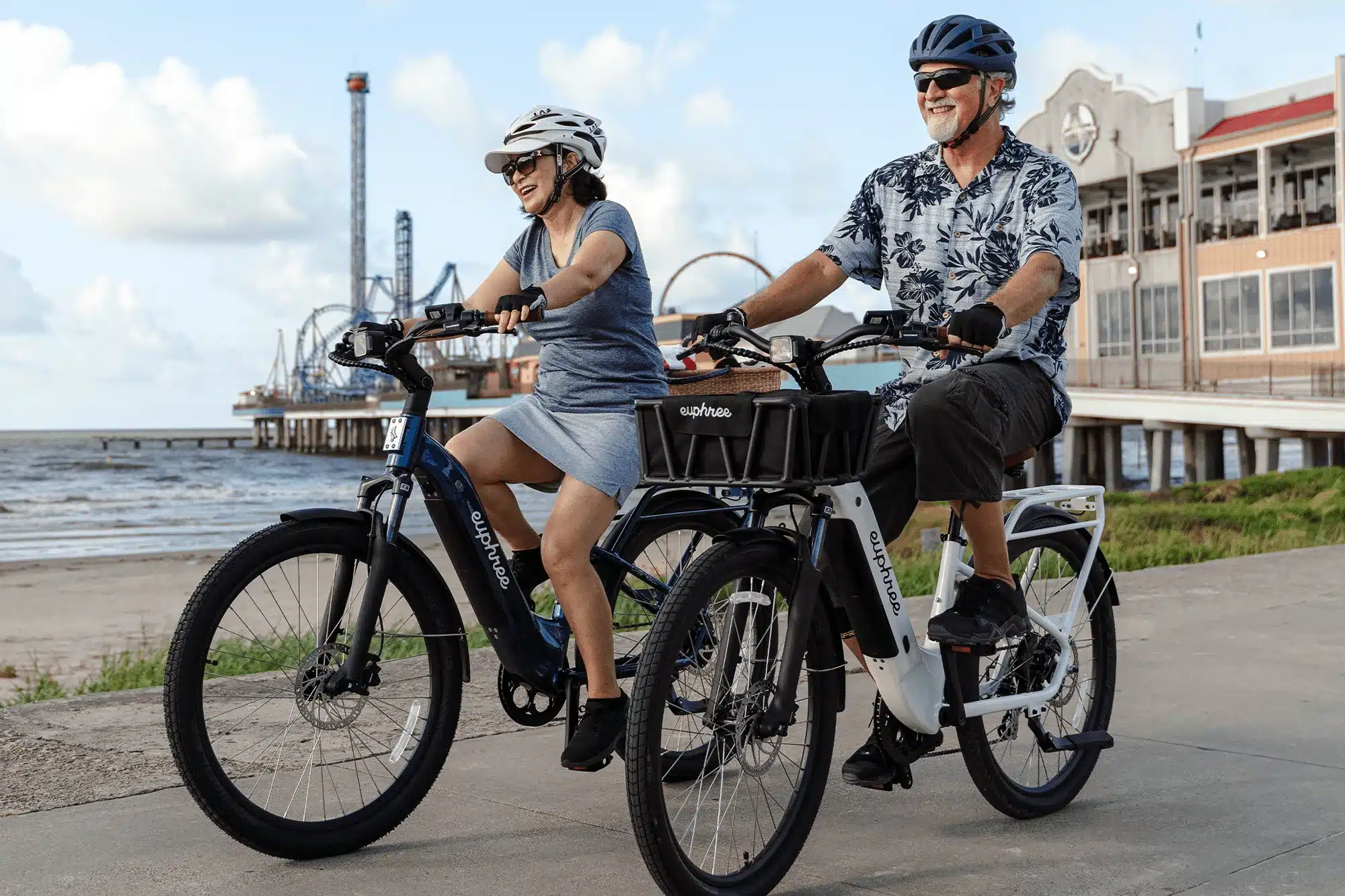 Ein Mann und eine Frau fahren nebeneinander auf zwei City Robin X+ Elektrofahrrädern am Strandweg. Im Hintergrund sind das Meer, ein Pier und Gebäude zu sehen.