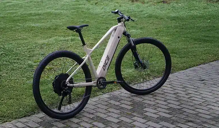 Ein hellbeiges Prophete E-MTB "Dice 2.0" mit 29-Zoll-Laufrädern und silbernem Rahmen steht auf einem gepflasterten Weg neben einer Rasenfläche.