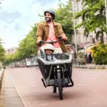 Ein Mann fährt ein Urban Arrow FamilyNext Lastenrad mit einem Kind im vorderen Kindersitz auf einer Straße in einer Stadt.