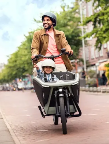 Ein Mann fährt ein Urban Arrow FamilyNext Lastenrad mit einem Kind im vorderen Kindersitz auf einer Straße in einer Stadt.