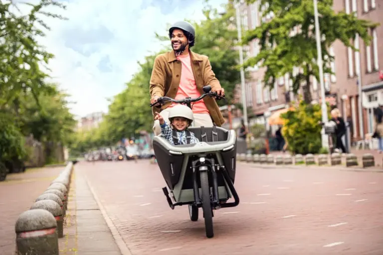 Ein Mann fährt ein Urban Arrow FamilyNext Lastenrad mit einem Kind im vorderen Kindersitz auf einer Straße in einer Stadt.