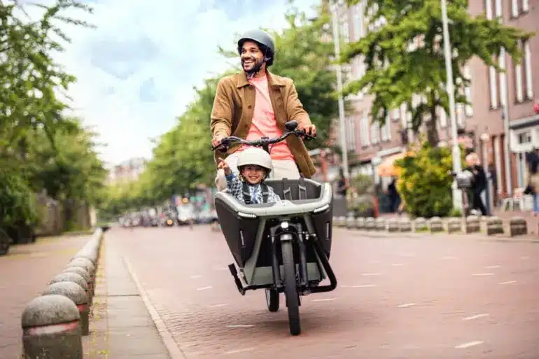 Ein Mann fährt ein Urban Arrow FamilyNext Lastenrad mit einem Kind im vorderen Kindersitz auf einer Straße in einer Stadt.