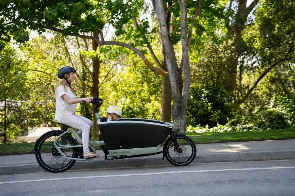 Elektrisches Lastenrad Urban Arrow FamilyNext mit Frau am Steuer und Kind in der Ladebox, fährt auf asphaltiertem Weg im Grünen.
