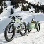 Ein hellgrünes E-Bike mit einem passenden Anhänger, in dem Skier transportiert werden, parkt auf einem schneebedeckten Hang mit Bäumen im Hintergrund.