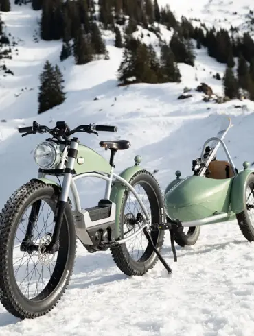 Ein hellgrünes E-Bike mit einem passenden Anhänger, in dem Skier transportiert werden, parkt auf einem schneebedeckten Hang mit Bäumen im Hintergrund.