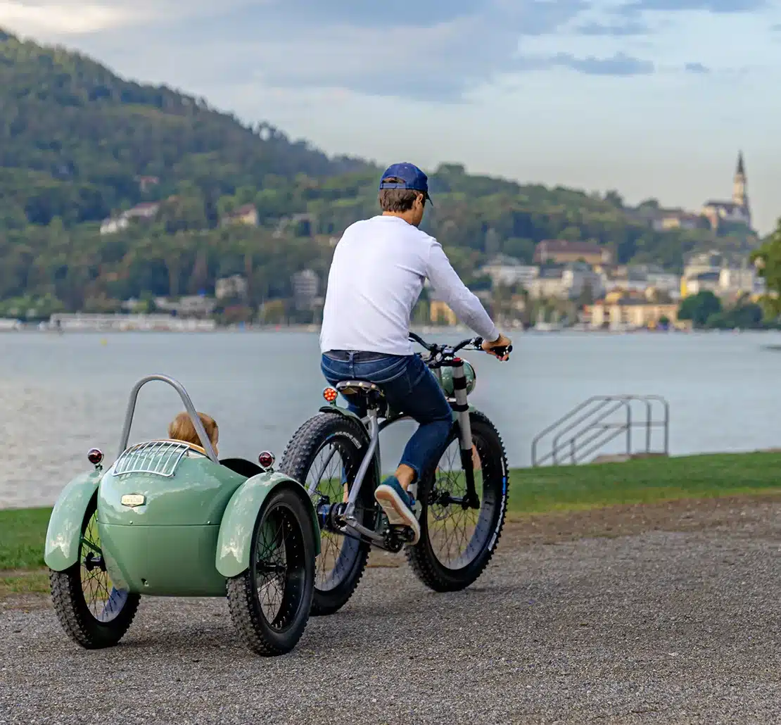 Ein Mann fährt mit einem Fahrrad, an das ein Kinderanhänger gekoppelt ist, am Ufer eines Sees entlang. Im Hintergrund sind Hügel mit Bäumen und einer Stadt zu sehen.