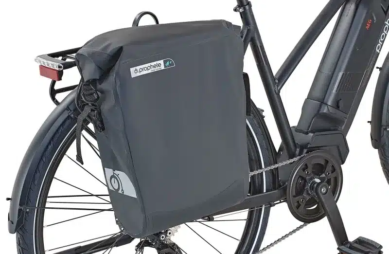 Seitenansicht eines schwarzen Prophete E-Trekkingrads mit einer großen, wasserdichten Gepäcktasche, die am Gepäckträger befestigt ist. Man sieht Teile des Hinterrads, des Rahmens, des Akkus und des Hinterradschutzes.