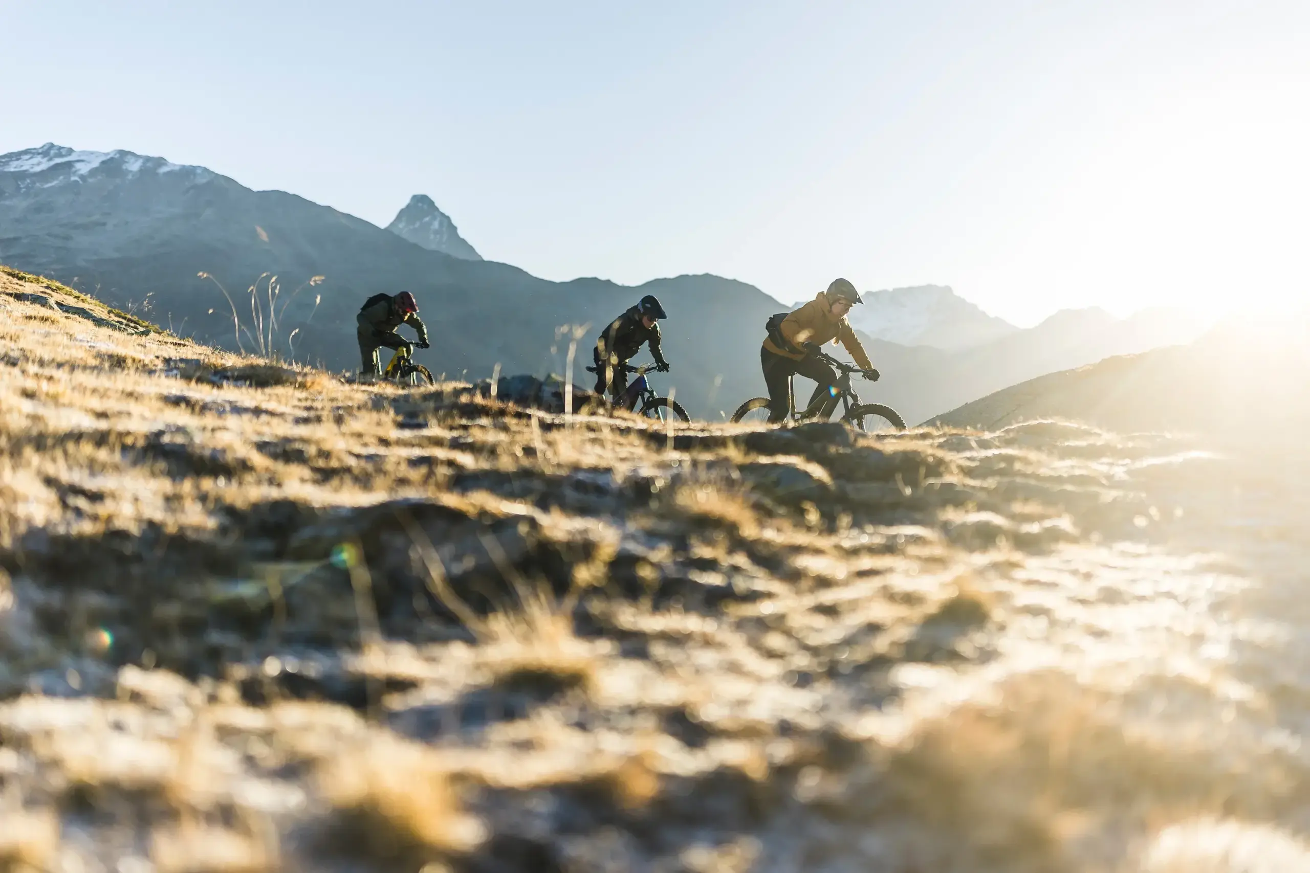 Drei Mountainbiker fahren mit ihren E-Bikes auf einem steinigen, grasbewachsenen Bergrücken bergab. Die Berge im Hintergrund sind schneebedeckt und die Sonne scheint von rechts.