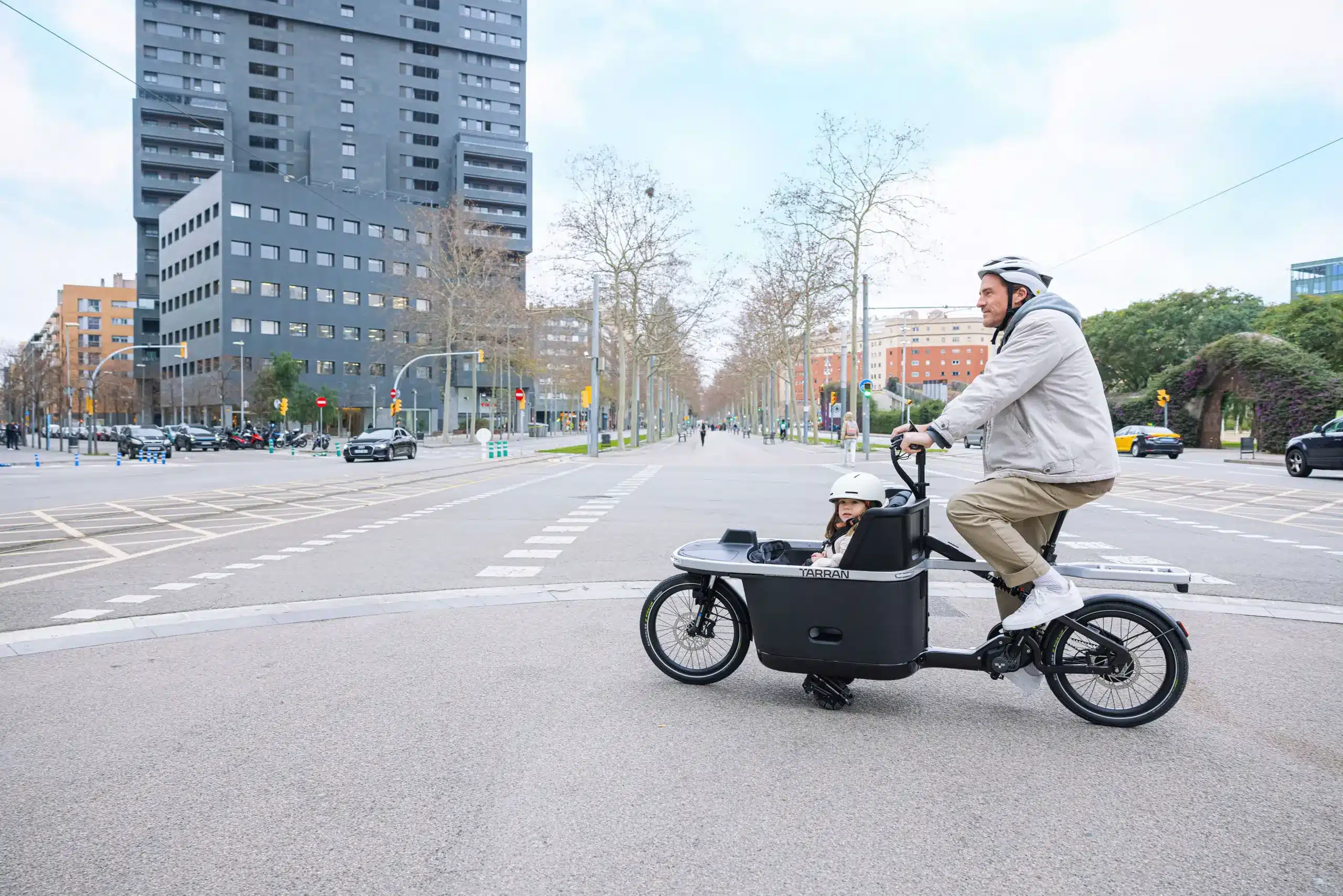 Ein Vater fährt mit seinem Kind in einem schwarzen Tarran T1 Pro Cargobike auf einer Straße in einer europäischen Stadt. Moderne Gebäude und Bäume säumen die Straße.