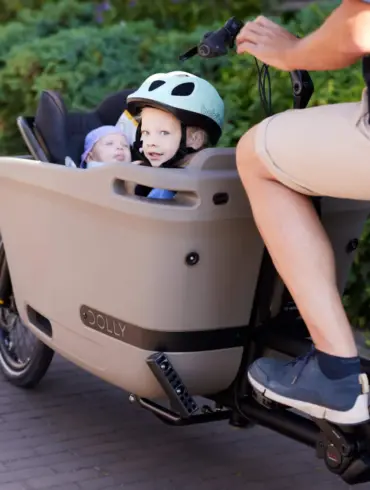 Kindertransport im Dolly Joy Lastenrad: Zwei Kinder sitzen in der beige farbenen Ladebox, während eine Person das Rad fährt. Das vordere Kind trägt einen mintgrünen Helm.