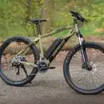 Ein olivgrünes E-Mountainbike der Marke "Graveler" steht seitlich auf einem Feldweg. Das Fahrrad hat breite Reifen, eine Federgabel und eine Batterie im Unterrohr.