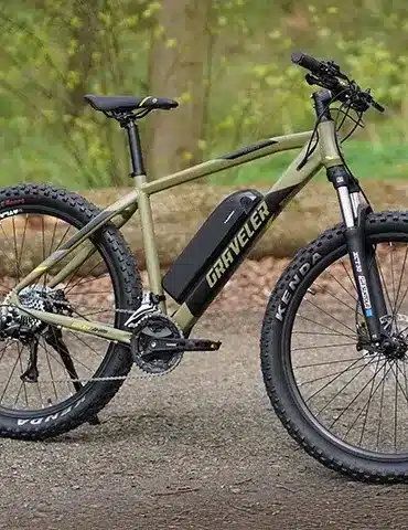 Ein olivgrünes E-Mountainbike der Marke "Graveler" steht seitlich auf einem Feldweg. Das Fahrrad hat breite Reifen, eine Federgabel und eine Batterie im Unterrohr.