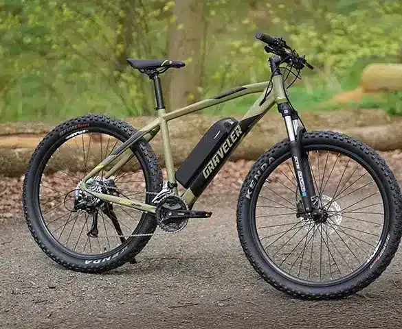 Ein olivgrünes E-Mountainbike der Marke "Graveler" steht seitlich auf einem Feldweg. Das Fahrrad hat breite Reifen, eine Federgabel und eine Batterie im Unterrohr.