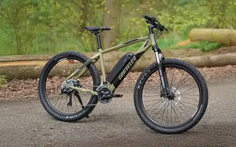 Ein olivgrünes E-Mountainbike der Marke "Graveler" steht seitlich auf einem Feldweg. Das Fahrrad hat breite Reifen, eine Federgabel und eine Batterie im Unterrohr.