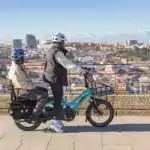 Ein blaues Tern GSD P00 E-Lastenrad fährt auf einer gepflasterten Straße. Eine Person fährt, eine andere sitzt auf einem Kindersitz im hinteren Bereich des Rads. Im Hintergrund ist eine Stadt mit Gebäuden und Hügeln zu sehen.