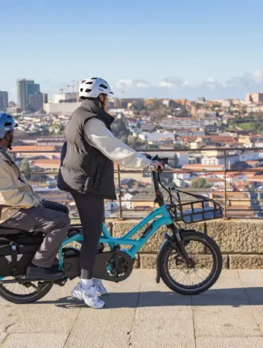 Ein blaues Tern GSD P00 E-Lastenrad fährt auf einer gepflasterten Straße. Eine Person fährt, eine andere sitzt auf einem Kindersitz im hinteren Bereich des Rads. Im Hintergrund ist eine Stadt mit Gebäuden und Hügeln zu sehen.