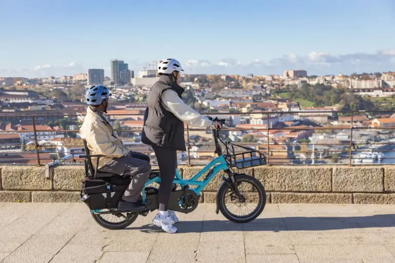 Ein blaues Tern GSD P00 E-Lastenrad fährt auf einer gepflasterten Straße. Eine Person fährt, eine andere sitzt auf einem Kindersitz im hinteren Bereich des Rads. Im Hintergrund ist eine Stadt mit Gebäuden und Hügeln zu sehen.