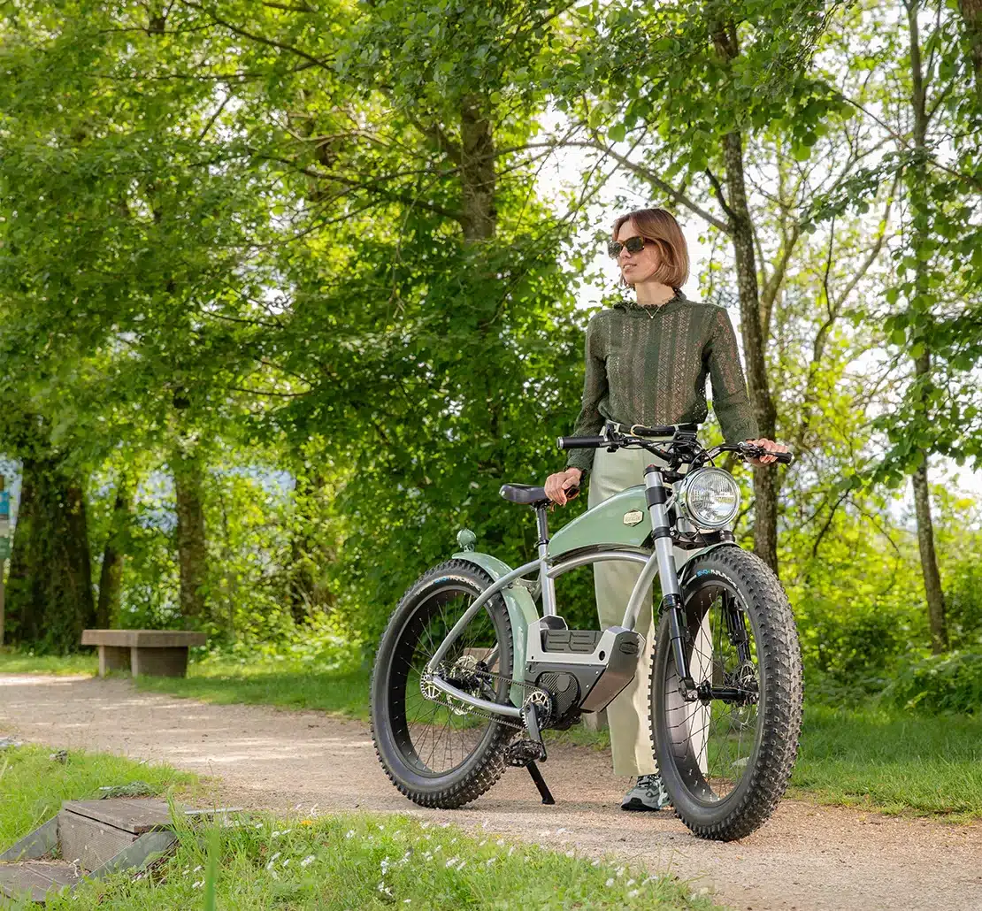 Eine junge Frau mit Sonnenbrille steht neben einem Heritage Origine E-Bike, das einen Stahlrahmen, breite Reifen und eine integrierte Motor-Akku-Einheit aufweist.
