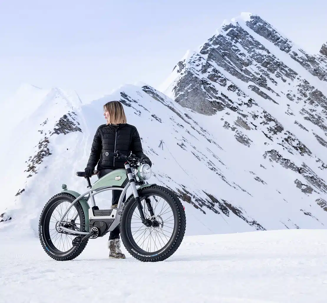 Frau mit einem mintgrünen E-Bike mit breiten Reifen und Vintage-Design in einer verschneiten Bergkulisse.