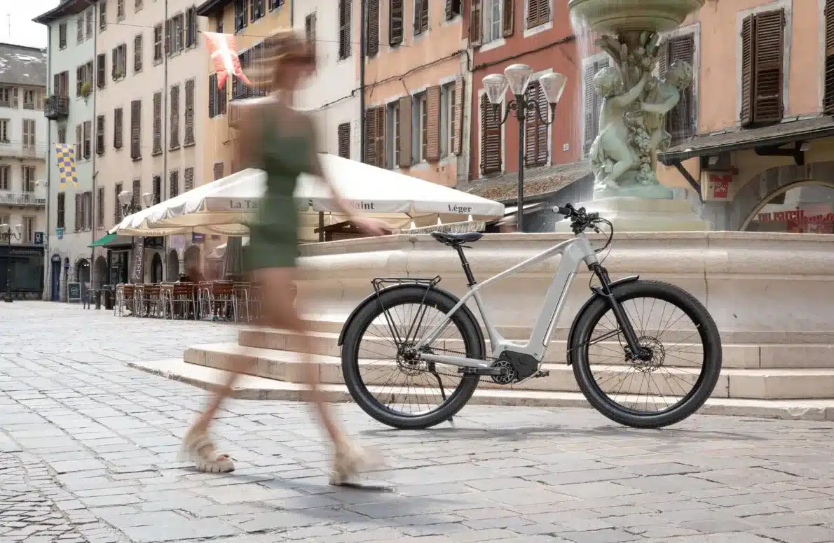 Graues Heritage Style E-Bike mit Gepäckträger und Schutzblechen steht auf einem Kopfsteinpflasterplatz vor einem Brunnen.