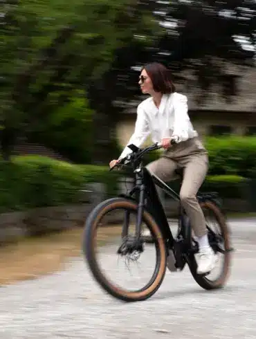 Eine Frau in stilvoller Kleidung fährt ein schwarzes E-Bike auf einem Schotterweg. Der Hintergrund ist unscharf und zeigt Bäume und ein Haus.