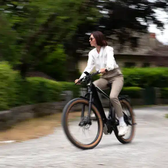 Eine Frau in stilvoller Kleidung fährt ein schwarzes E-Bike auf einem Schotterweg. Der Hintergrund ist unscharf und zeigt Bäume und ein Haus.