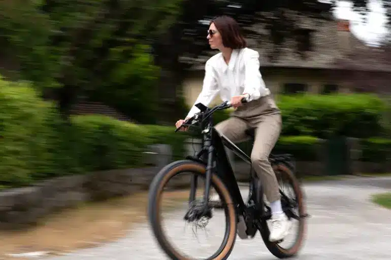 Eine Frau in stilvoller Kleidung fährt ein schwarzes E-Bike auf einem Schotterweg. Der Hintergrund ist unscharf und zeigt Bäume und ein Haus.